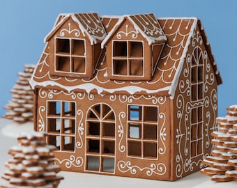 May include: A brown gingerbread house with white icing details, featuring two dormer windows on the roof, a central arched doorway, and multiple square window cutouts. The structure is decorated with white piped scrollwork and roof trim.