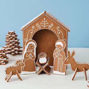May include: A gingerbread nativity scene featuring a stable structure with white icing details, figures of two adults, a baby in a manger, a sheep, and a donkey. The set includes two stacked gingerbread trees with white icing accents on the edges.