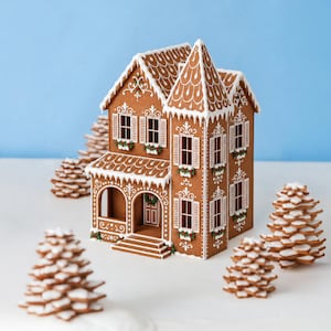 May include: A brown gingerbread house with white icing details, including roof shingles, window shutters, and floral accents. The structure features a front porch with steps and is surrounded by several small, stacked gingerbread pine trees.
