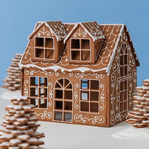 Può includere: Una casa di pan di zenzero con dettagli di glassa bianca e alberi di pan di zenzero decorativi. La casa ha più finestre e abbaini, ed è ambientata su uno sfondo azzurro. Una decorazione festiva per le vacanze.