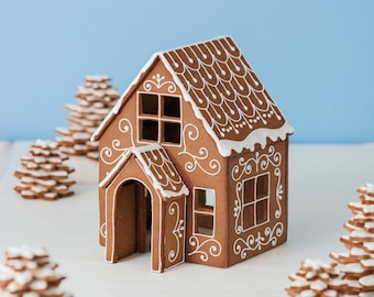 May include: A brown gingerbread house featuring white icing details in a scalloped roof pattern and swirling scrollwork designs on the walls. The structure includes a front porch, open windows, and a doorway, surrounded by small cookie trees.