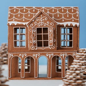 May include: A brown gingerbread house featuring white icing details on the roof, walls, and window frames. The structure has a two-story design with an arched ground floor and a central dormer window, decorated with intricate white piping patterns.