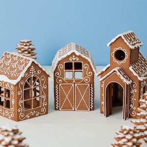 May include: Three brown gingerbread house structures decorated with white icing patterns. The set includes a house with arched windows, a barn with double doors, and a church with a steeple. Several small stacked gingerbread trees are also present.