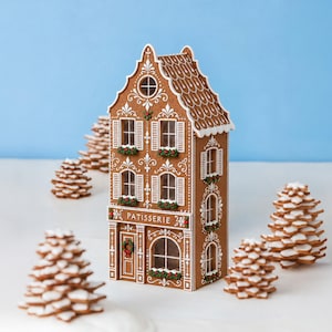 Könnte beinhalten: Ein Lebkuchenhaus-Modell mit der Aufschrift PATISSERIE, weißen Zuckerguss-Details, Bogenfenstern und Blumenkästen, umgeben von mehreren kleinen, gestapelten Lebkuchen-Tannenbäumen mit weißem Zuckerguss auf einer weißen Fläche vor blauem Hintergrund.