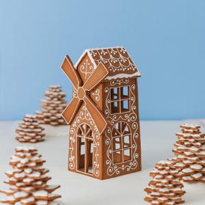 May include: A gingerbread windmill house with white icing details and a windmill. The house has windows and decorative icing patterns. Several gingerbread tree-shaped biscuits are in the background, all on a white surface against a blue backdrop.