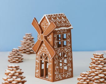 May include: A gingerbread windmill house with white icing details and a windmill. The house has windows and decorative icing patterns. Several gingerbread tree-shaped cookies are in the background, all on a white surface against a blue backdrop.