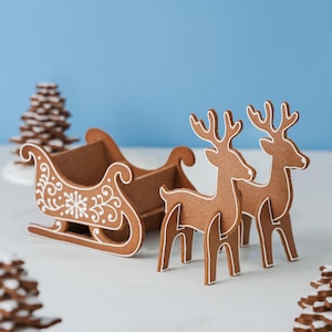 Puede incluir: Decoraciones navideñas de pan de jengibre. Un trineo marrón con detalles de glaseado blanco y dos renos son el punto focal. Los renos y el trineo están sobre una superficie blanca con un fondo azul. También se ven árboles de pan de jengibre.