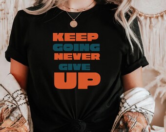 May include: Black t-shirt with the words "KEEP GOING NEVER GIVE UP" in orange and blue block letters. The shirt is knotted at the waist and has rolled-up sleeves. A patterned jacket is draped over the shoulders.