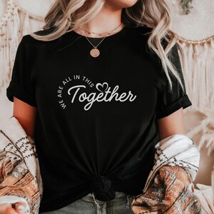 Puede incluir: Camiseta negra con el texto blanco "WE ARE ALL IN THIS Together" en una fuente cursiva. La palabra "Together" tiene un símbolo de corazón que reemplaza la letra "o". La camiseta está anudada en la cintura.