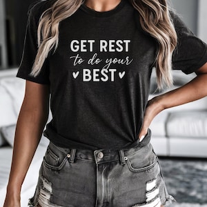 Puede incluir: Camiseta gris oscuro con el texto "GET REST to do your BEST" en escritura blanca. La camiseta se combina con shorts de mezclilla desgastados. El estilo general es casual y relajado.
