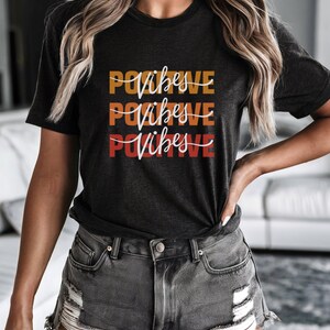 May include: Dark gray t-shirt with the words "Positive Vibes" in a stacked design. The text is in a gradient of orange and red. The shirt is paired with gray denim shorts.