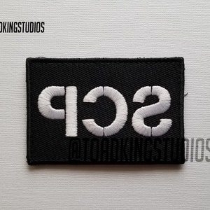 以下が含まれることがあります： 黒い刺繍パッチに白い文字で「SC1F」と書いてあります。