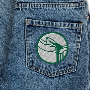 以下が含まれることがあります： ライトブルーのデニムジーンズ。バックポケットには円形のパッチが付いています。パッチは白い背景に、手と流れるラインの緑色のデザインが施されています。ジーンズは折りたたまれており、デニムの質感がわかります。