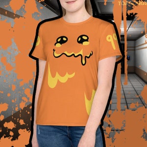 以下が含まれることがあります： オレンジ色のTシャツに、漫画の顔のグラフィックがプリントされています。顔は黄色で、目は黒く、口は垂れています。Tシャツの右側に99とあります。