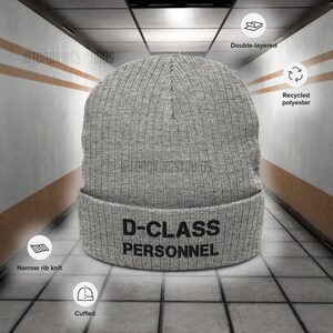 May include: Grey knitted beanie with a black embroidered design that reads "D-CLASS PERSONNEL". The beanie is double-layered, made from recycled polyester, and has a narrow rib knit cuff.