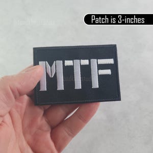 K&ouml;nnte beinhalten: Schwarzer rechteckiger Aufn&auml;her mit wei&szlig; gestickten Buchstaben "MTF". Der Aufn&auml;her ist 7,6 cm gro&szlig;. Der Aufn&auml;her wird in einer Hand gehalten.