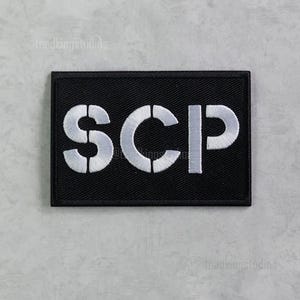 Puede incluir: Parche rectangular negro con las letras SCP bordadas en blanco. El parche tiene un borde negro. Las letras están en una fuente en negrita sin serifa.