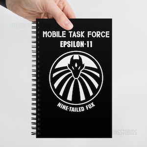 Puede incluir: Cuaderno negro de espiral con un logotipo blanco que dice "Mobile Task Force Epsilon-11 Nine-Tailed Fox". El logotipo presenta una cabeza de zorro estilizada con un círculo de líneas radiantes.
