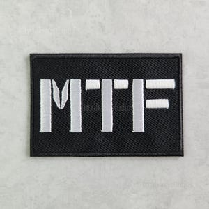 K&ouml;nnte beinhalten: Schwarzer rechteckiger Aufn&auml;her mit den wei&szlig; gestickten Buchstaben "MTF". Der Aufn&auml;her hat einen schwarzen Rand und ist wahrscheinlich zum Aufn&auml;hen oder Befestigen an Kleidung oder Ausr&uuml;stung gedacht. Die Buchstaben sind in einer fetten, serifenlosen Schrift.