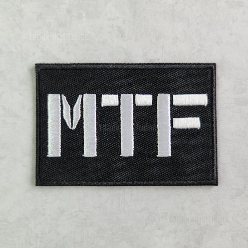 K&ouml;nnte beinhalten: Schwarzer rechteckiger Aufn&auml;her mit den wei&szlig; gestickten Buchstaben "MTF". Der Aufn&auml;her hat einen schwarzen Rand und ist wahrscheinlich zum Aufn&auml;hen oder Befestigen an Kleidung oder Ausr&uuml;stung gedacht. Die Buchstaben sind in einer fetten, serifenlosen Schrift.