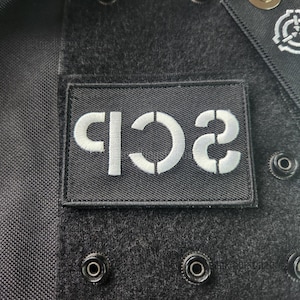 Puede incluir: Primer plano de una tela negra con un parche rectangular bordado con las letras "C B 2" en blanco. La tela tiene varios cierres de presión metálicos y un parche más pequeño con un símbolo.