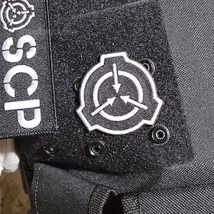 May include: Black tactical vest with white SCP logo patches. The patches feature a circular design with arrows and the letters "SCP". The vest has multiple pockets and straps.