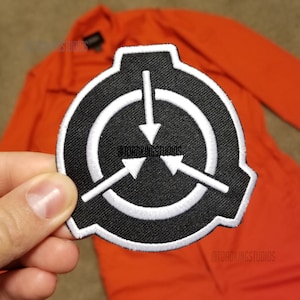 May include: A black and white embroidered patch featuring a circular design with three arrows pointing inward. The patch is held in front of an orange garment. The patch has a white border and white stitching.
