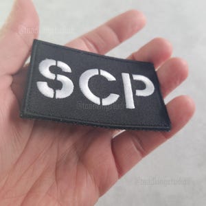 May include: Black rectangular patch with the letters "SCP" in white, stencil-style font. The patch is held in a hand against a light background. The text is centered on the patch.