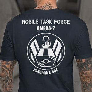 Puede incluir: Una camiseta azul marino con un gráfico blanco de un círculo que contiene una caja con un ojo, una mano y el texto "Pandora's Box". El texto "Mobile Task Force Omega-7" está por encima del gráfico.