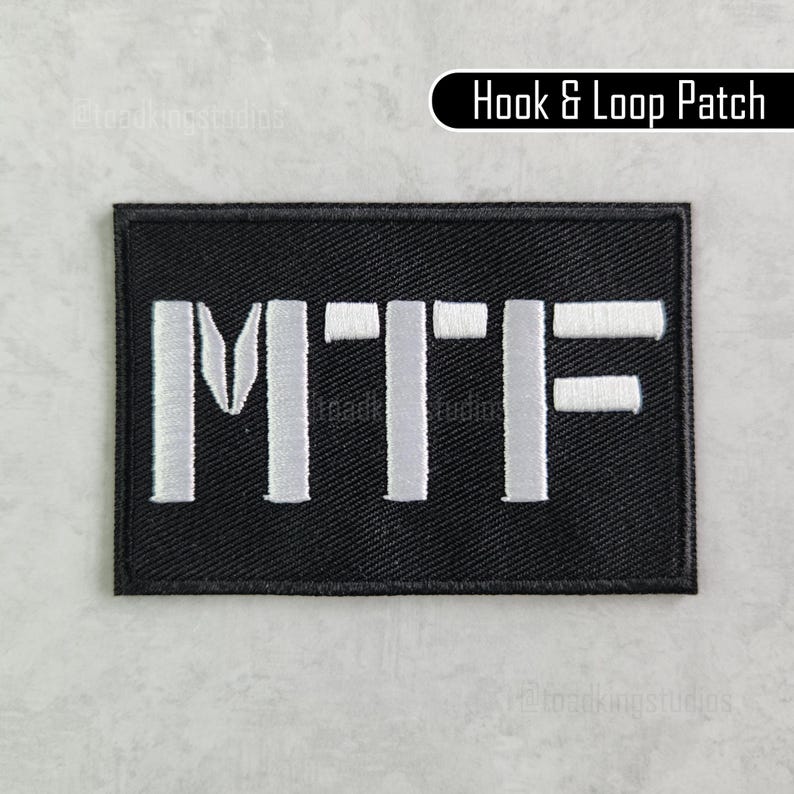 K&ouml;nnte beinhalten: Ein schwarzer rechteckiger Klettverschluss-Patch mit den Buchstaben "MTF" in Wei&szlig; gestickt. Der Patch hat einen schwarzen Rand und den Text "Hook & Loop Patch" in wei&szlig;er Schrift auf schwarzem Hintergrund.