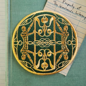 May include: A round, embroidered patch with a dark green background and gold-colored detailing. The design features two stylized figures holding up a central structure, with the text "Property of The Wanderer's Library" on a library card.