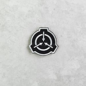 May include: A black and white embroidered patch featuring the SCP Foundation logo. The logo is a stylized shield shape with a central circle containing three arrows pointing inward. The patch has a white border.