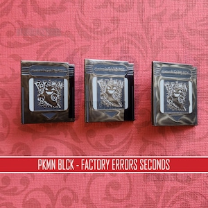 May include: Three vintage Nintendo Game Boy cartridges. Each cartridge features a metallic finish and a Pokémon graphic. The text "PKMN BLCK - FACTORY ERRORS SECONDS" is displayed on a red banner below the cartridges.