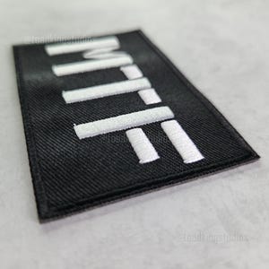 K&ouml;nnte beinhalten: Schwarzer rechteckiger Aufn&auml;her mit wei&szlig; gesticktem Text "MTTF". Der Aufn&auml;her hat eine strukturierte Oberfl&auml;che und ist leicht angewinkelt, wodurch die wei&szlig;en N&auml;hte vor dem dunklen Hintergrund hervorgehoben werden. Es ist wahrscheinlich ein Accessoire.