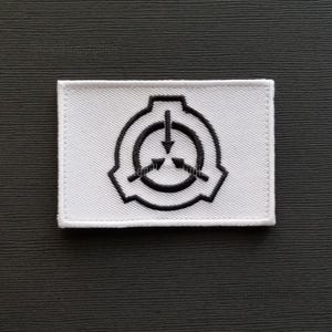 May include: A white rectangular patch with a black emblem featuring a circle with an arrow pointing down and three arrows pointing inward. The patch is on a dark grey textured surface.