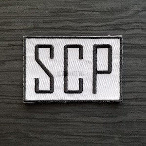以下が含まれることがあります： 白い長方形のパッチに、黒い刺繍で「SCP」と綴られています。