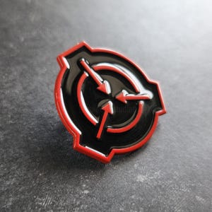 May include: A circular metal enamel pin featuring a black center with a red border. The design displays a stylized emblem with three red arrows pointing inward toward the center, enclosed within a circular frame that has a notched outer edge.