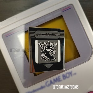 May include: A silver-colored enamel pin featuring a Game Boy cartridge with the text "Nintendo GAME BOY" and a pixelated image of the Pokemon Ghost-type, Gengar. The pin is designed to look like a Game Boy cartridge.