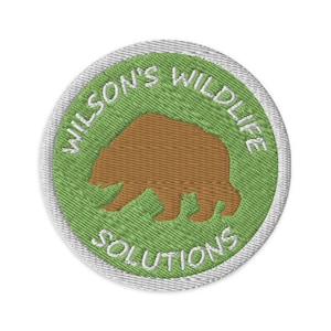 May include: Embroidered patch with a brown bear silhouette on a green background. The text "WILSON'S WILDLIFE SOLUTIONS" is embroidered in white around the bear. The patch has a white border.