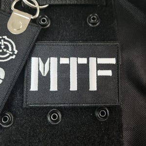 K&ouml;nnte beinhalten: Nahaufnahme eines schwarzen Stoffes mit einem wei&szlig; gestickten "MTF"-Patch. Der rechteckige Patch ist mittig platziert. Silberne Metallbeschl&auml;ge und ein kleines Etikett mit wei&szlig;em Logo sind sichtbar. Mehrere schwarze Druckkn&ouml;pfe sind ebenfalls vorhanden.