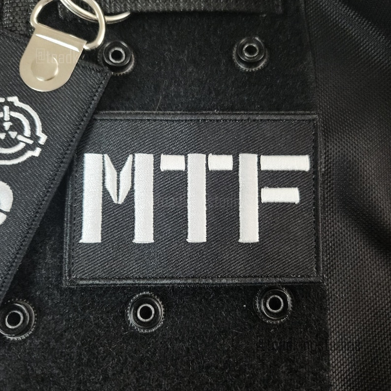K&ouml;nnte beinhalten: Nahaufnahme eines schwarzen Stoffes mit einem wei&szlig; gestickten "MTF"-Patch. Der rechteckige Patch ist mittig platziert. Silberne Metallbeschl&auml;ge und ein kleines Etikett mit wei&szlig;em Logo sind sichtbar. Mehrere schwarze Druckkn&ouml;pfe sind ebenfalls vorhanden.