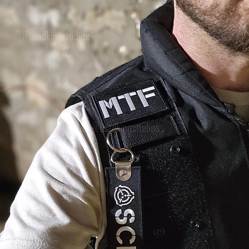 K&ouml;nnte beinhalten: Schwarze taktische Weste mit den Buchstaben "MTF" auf einem Aufn&auml;her. Ein Schl&uuml;sselband mit dem SCP-Logo und Text h&auml;ngt an der Weste. Die Weste wird &uuml;ber einem hellfarbigen Hemd getragen.