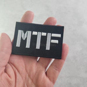 K&ouml;nnte beinhalten: Schwarzer rechteckiger Aufn&auml;her mit den Buchstaben "MTF" in Wei&szlig; gestickt. Der Aufn&auml;her wird in einer Hand gehalten. Der Text ist auf dem Aufn&auml;her zentriert.