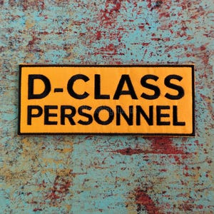 May include: A rectangular yellow patch with black embroidered text that reads "D-CLASS PERSONNEL". The patch has a black border and is set against a textured, multi-colored background.