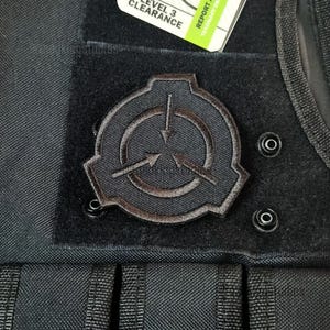May include: Black patch featuring the SCP Foundation logo, a stylized circle with three arrows pointing inward. The patch is sewn onto a black fabric, with a tag that reads "Level 3 Clearance".
