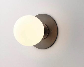 May include: A modern wall sconce featuring a round, frosted glass globe emitting a soft glow. The globe is mounted on a circular, dark bronze-colored metal backplate. The fixture is designed for indoor lighting.