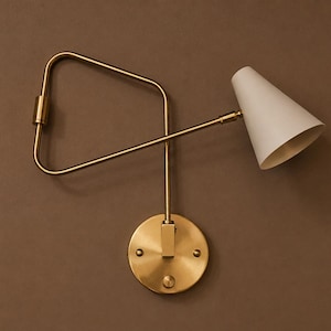 Peut inclure: Une applique murale avec une finition en laiton brossé, une plaque de base circulaire et une structure de bras réglable. La lampe comprend un abat-jour conique blanc cassé fixé à l'extrémité du bras, offrant une source de lumière directionnelle.