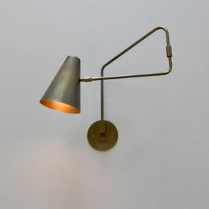 Op de afbeelding: Een messing wandlamp met een conische kap en een verstelbare arm. De kap heeft een warme, gouden binnenkant en de armatuur is gemonteerd op een cirkelvormige basis. Het ontwerp is minimalistisch en modern.