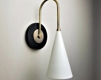 May include: A modern wall sconce with a black circular base and a brass-colored curved arm. The sconce features a white, cone-shaped lampshade. The design is minimalist and elegant, suitable for various interior styles.