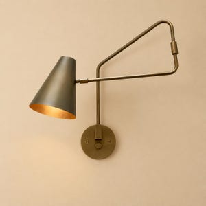 May include: A wall-mounted lamp featuring a conical shade with a dark exterior and a warm metallic interior. The fixture includes an adjustable arm with a triangular support frame, all finished in a muted brass tone, attached to a circular base.